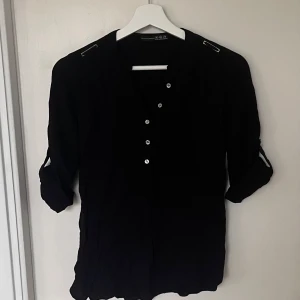 Svart blus med knappar - Elegant svart blus med knappar framtill och dekorativa axeldetaljer. Blusen har trekvartsärmar med knappdetalj vid ärmslut. Perfekt för en stilren look.