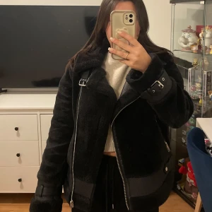 Svart teddyjacka med dragkedja - Säljer en snygg svart teddyjacka med dragkedja och spänndetaljer. Jackan har en mysig krage och långa ärmar. Perfekt för kyliga dagar!