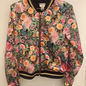 Blommig bomberjacka från H&M - Snygg och färgglad bomberjacka från H&M i samarbete med Angela McKay. Jackan har ett blommigt mönster i rosa, gult och grönt på en mörk bas. Den har dragkedja framtill och ribbade muddar vid ärmslut och nederkant.