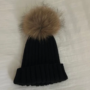Svart ribbad mössa med pälsboll - Snygg svart ribbad mössa med en stor beige pälsboll på toppen. Perfekt för att hålla värmen under kalla dagar. Stilren och enkel design som passar till det mesta.