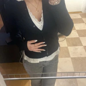 Svart kofta med spetsdetaljer - Säljer en stilren svart kofta med knappar framtill. Koftan har långa ärmar och en vacker spetskant vid halsringningen som ger en elegant touch. Perfekt för lager-på-lager-stil.