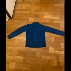 Blå fleecejacka från Arc'teryx - Säljer en blå fleecejacka från Arc'teryx med dragkedja framtill. Perfekt för kyliga dagar och outdoor-äventyr. Jackan har långa ärmar och en stilren design.