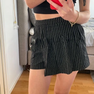 Svart randig omlottkjol - Trendig svart omlottkjol med vita ränder och praktiska fickor. Kjolen har en D-ring detalj och elastisk midja och inbyggda shorts!