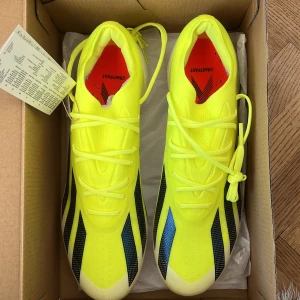 Adidas X Crazyfast Elite FG Fotbollsskor - Säljer ett par Adidas X Crazyfast Elite FG fotbollsskor i en iögonfallande gul färg med svarta och blå detaljer. Skorna har snörning och är designade för att ge optimal prestanda på fotbollsplanen. Perfekta för spel på fasta gräsplaner.