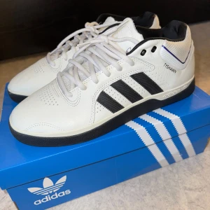 Adidas Tyshawn  - Snygga vita Adidas Tyshawn skor med svarta detaljer och klassiska tre ränder. Perfekta för skate eller vardagsbruk. Skorna har en bekväm passform.