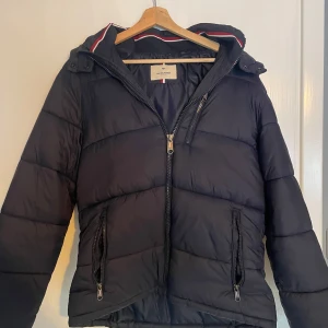 Svart pufferjacka från Jack & Jones - Snygg svart pufferjacka från Jack & Jones med huva och dragkedja. Jackan har en stilren design med ribbade detaljer och ett märke på ärmen. Perfekt för kyliga dagar.