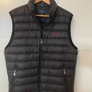 Svart dunväst från Polo Ralph Lauren - Snygg svart dunväst från Polo Ralph Lauren med dragkedja och broderad logga i rött. Perfekt för lager-på-lager under kyliga dagar.