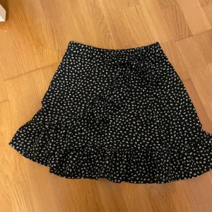 Svart blommig kjol från FB Sister - Säljer en superfin svart kjol med små vita blommor från FB Sister. Kjolen har en söt volangdetalj längst ner och en knytning i midjan som ger en extra touch. Perfekt för både vardag och fest!