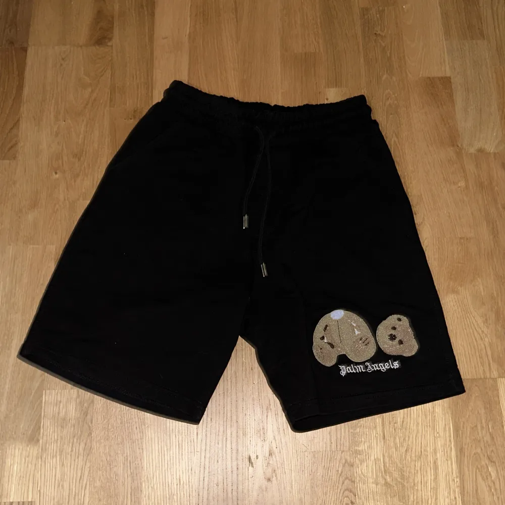 Säljer ett par svarta shorts från Palm Angels. De är i bra skick och har en cool design med en nallebrodyr på framsidan. Shortsen har en elastisk midja med dragsko för bästa passform. Perfekta för en avslappnad stil!. Shorts.