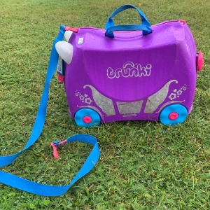 Lila resväska från Trunki - Säljer en superfin lila resväska från Trunki. Den har en rolig design med blå hjul och handtag, samt vita detaljer som ser ut som horn. Perfekt för barn att sitta på och rulla runt med. Väskan har en dragkedja och en blå axelrem för enkel transport.