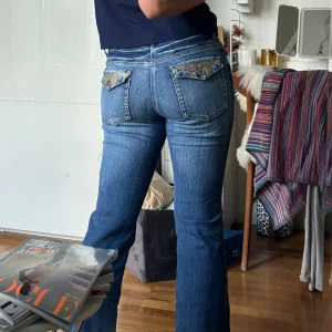 Blå jeans med broderade fickor - Säljer ett par snygga blå jeans med bootcut-stil. De har coola broderade detaljer på bakfickorna som verkligen sticker ut. Perfekta för en avslappnad look men med en touch av stil. Passar bra till både vardag och fest!