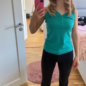 Turkos ribbad linne - Säljer en snygg turkos ribbad linne med spetsdetaljer vid axelbanden och knappar framtill. Perfekt för sommardagar eller som ett lager under en kofta. Linnet är i bra skick och superbekvämt att ha på sig.