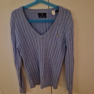 Ljusblå stickad tröja från Gant - Säljer en ljusblå stickad tröja från Gant med kabelstickat mönster och v-ringning. Tröjan har långa ärmar och en liten logga på bröstet. Perfekt för kyliga dagar!