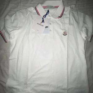 Vit Moncler T-shirt - Säljer en stilren vit pikétröja från Moncler med klassisk krage och knappar framtill. Tröjan har Monclers logotyp broderad på bröstet och röd-blå ränder på krage och ärmslut. Perfekt för en avslappnad men ändå elegant look.