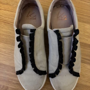 Beige sneakers med svarta volanger från K Cobbler - Snygga beige sneakers från K Cobbler med unika svarta volanger längs ovansidan. Perfekta för en stilren och trendig look. Skorna har en bekväm passform och är lätta att matcha med olika outfits.