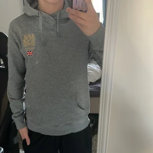 Grå hoodie från Morris - Säljer en stilren grå hoodie från Morris med broderad logga och brittisk flagga på bröstet. Tröjan har en klassisk känguruficka och justerbar huva med dragsko. Perfekt för en avslappnad stil. Pris kan diskuteras 