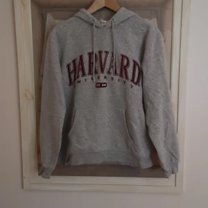 Grå hoodie från H&M med Harvard-tryck - Säljer en grå hoodie från H&M med stort Harvard University-tryck i vinrött på framsidan. Tröjan har en klassisk känguruficka och justerbar huva med dragsko. 