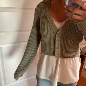 Grön kofta - Säljer en grön ribbad kofta från Divided. Den är croppad med knappar framtill och har långa ärmar. Perfekt för lager-på-lager-stil!