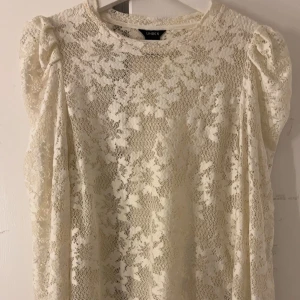 Vit spetsblus från Lindex - Elegant vit spetsblus från Lindex med korta ärmar och ett vackert blommönster. Perfekt för en stilren look med en romantisk touch. Blusen har en rund halsringning och är tillverkad i ett lätt och luftigt material. Storlek M men sitter som en S