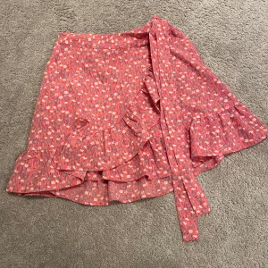 Röd blommig omlottkjol - Charmig röd omlottkjol med blommigt mönster och volangdetaljer. Perfekt för en somrig look med sin lätta och luftiga design.