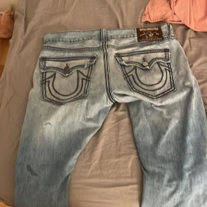 Ljusa slitna true religion - Säljer ett par ljusa trues med slitningar och broderade detaljer på bakfickorna. Jeansen har en loose passform och är perfekta för en avslappnad stil. De har en klassisk femficksdesign och knappgylf.