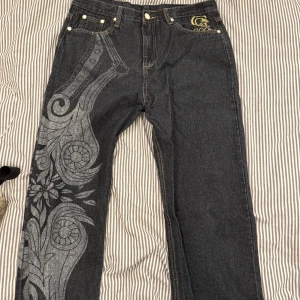 Unika jeans med broderi från Coogi - Snygga jeans från Coogi med unikt broderi i rött och guld på bakfickorna. De har ett stort, dekorativt mönster längs ena benet och klassisk femficksdesign. Perfekta för dig som vill sticka ut!