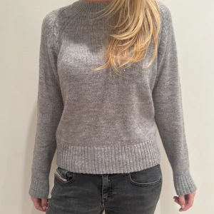 Grå stickad tröja från Indiska  - Säljer en mysig grå stickad tröja med flätad detalj längs ärmarna. Tröjan har långa ärmar och ribbstickade muddar vid ärmslut och nederkant. Perfekt för kyliga dagar!