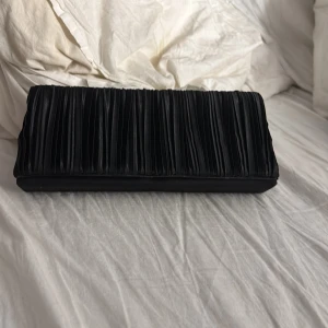 Svart kuvertväska med veckad design - Elegant svart kuvertväska med en unik veckad design. Perfekt för att ge en stilren touch till din outfit. Väskan är kompakt och lätt att bära med sig.
