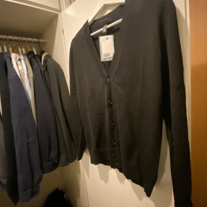 Svart cardigan/kofta från H&M - Säljer en stilren svart cardigan från H&M med knappar framtill. Koftan har en v-ringad design och långa ärmar, perfekt för lager-på-lager under kyligare dagar. Tröja är helt oanvänd som ni ser så är tagen även kvar! Storlek XS, men väldigt stor i sina storlek! Så passar garanterat S och någon som är slim M