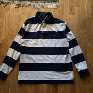 Randig pikétröja från Gant - Säljer en klassisk randig pikétröja från Gant i marinblått och vitt. Tröjan har långa ärmar och en stilren krage med knappar. Perfekt för en avslappnad men ändå stilfull look.