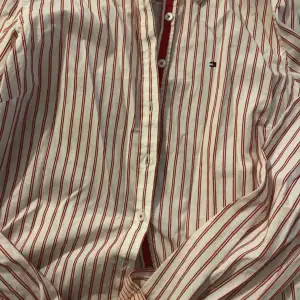 Snygg vit och röd randig skjorta från Tommy Hilfiger. Skjortan har en klassisk krage och knappar framtill. Perfekt för en stilren look.