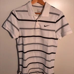 Vit och grå randig pikétröja från Nike - Snygg vit pikétröja från Nike med grå och svarta ränder. Tröjan har korta ärmar och en klassisk krage med knappar. Perfekt för en sportig och avslappnad stil. Knappt använd. Pris kan diskuteras
