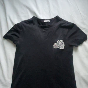 Svart moncler t-shirt - Snygg svart moncler t-shirt med ett coolt tryck på bröstet. Perfekt för en avslappnad stil. Kortärmad och i mjukt material.