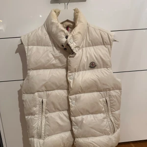 Vit dunväst från Moncler - Säljer en stilren vit dunväst från Moncler med hög krage och knappar framtill. Västen har två praktiska fickor med dragkedja och Moncler-loggan på bröstet. Perfekt för kyliga dagar! ny pris:5000