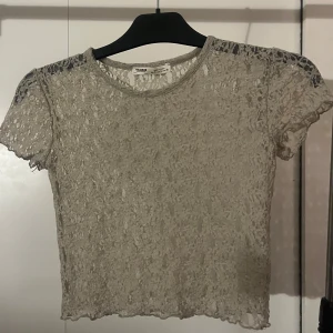 Grå spetsblus från Pull&Bear - Säljer en elegant grå spetsblus från Pull&Bear. Blusen har korta ärmar och ett vackert spetsmönster som ger en romantisk touch. Perfekt för att lägga till lite stil i din garderob.
