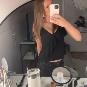 Svart volanglinne - Snyggt svart linne med v-ringad design och volangdetaljer. Perfekt för en stilren look. Den har axelband ❤️