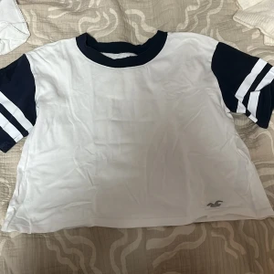 Vit och marinblå t-shirt från Hollister - Säljer en vit och marinblå t-shirt från Hollister med korta ärmar och randiga detaljer. T-shirten har en rund halsringning och är i en croppad stil. Perfekt för en avslappnad look.