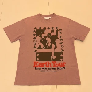 Rosa/ljus lila t-shirt från Butter - Lila t-shirt från Butter med ett brungrått tryck på framsidan. T-shirten har korta ärmar och en avslappnad passform. Skriv om ni har frågor.