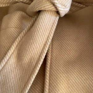 Beige kappa med knytband - Snygg beige kappa med knytband i midjan och stora fickor framtill. Perfekt för höst och vår, denna kappa har en stilren design utan krage och är tillverkad i ett mjukt material.