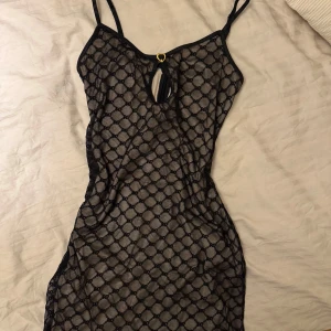 Svart mönstrad klänning med hjärtdetalj - Snygg svart klänning med ett genomskinligt mönster och en söt hjärtdetalj framtill. Klänningen har smala axelband och en tight passform som framhäver figuren. Perfekt för en utekväll eller speciella tillfällen.
