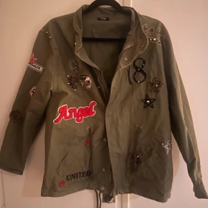 Grön parkas - Säljer en grön jacka med coola patches och broderier, inklusive texten 'Angel'. Jackan har en avslappnad passform och knappar framtill. Perfekt för en unik stil!