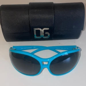 Blå solglasögon från Dolce & Gabbana - Snygga blå solglasögon från Dolce & Gabbana med svarta linser och en stilren design. Glasögonen har en bred båge och D&G-logga på sidan. Perfekta för att ge en färgklick till din outfit.