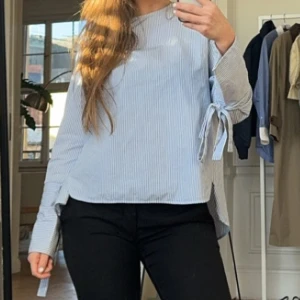 Randig blus med knytband - Snygg och stilren randig blus i ljusblått och vitt. Blusen har långa ärmar och knytband vid ärmsluten för en extra detalj. Perfekt för en chic och avslappnad look. 