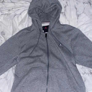 Grå hoodie från Jordan - Säljer en stilren grå hoodie från Jordan med dragkedja och den ikoniska loggan på bröstet. Perfekt för en avslappnad look. Den har en justerbar huva och är tillverkad i mjukt material för extra komfort.
