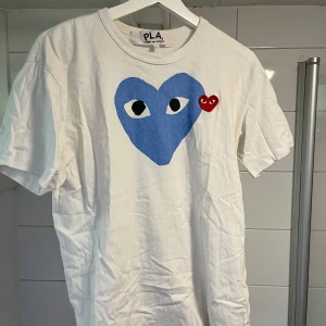 Vit t-shirt från Comme des Garçons - Säljer en vit t-shirt från Comme des Garçons med ett stort blått hjärta och ett litet rött hjärta med ögon på framsidan. T-shirten är kortärmad och i bomull, perfekt för en stilren look. Nypris ligger på ungefär 1000 kr. Tröjan är i L men passar även M, då den är lite mindre i storlek. Köpt på NK i Stockholm. Tröjan är skrynklig på bild då den legat i garderoben länge.