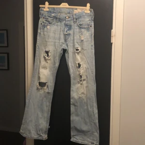 Ljusa slitna jeans från Hollister - Säljer ett par ljusa jeans från Hollister med slitna detaljer och hål för en trendig look. De har en klassisk femficksdesign och knappgylf. Perfekta för en avslappnad stil.