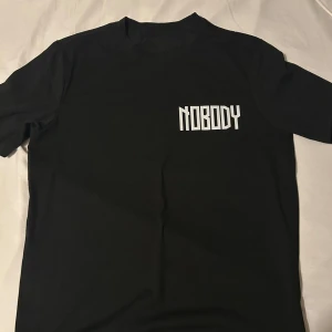 Svart t-shirt med tryck - Cool svart t-shirt med texten 'NOBODY' på framsidan och ett stort tryck med texten 'TRUST NOBODY' och en mask på baksidan. Perfekt för en edgy look.