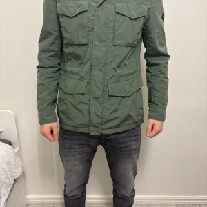 Grön jacka med fickor - Snygg grön jacka med flera praktiska fickor och en logga på ärmen. Jackan har långa ärmar och en stilren design som passar perfekt för höst och vår. Den är tillverkad i ett slitstarkt material och har en normal passform. Den är nästan aldrig använd max 1-2 gånger. Det står M men har själv s och den passar mig 