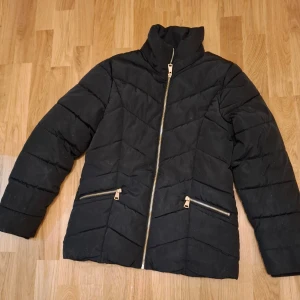 Svart pufferjacka från KappAhl - Snygg svart pufferjacka från KappAhl med dragkedja och två fickor framtill. Jackan har en hög krage och quiltad design som ger extra värme. Perfekt för kyliga dagar. Nypris 600 kr