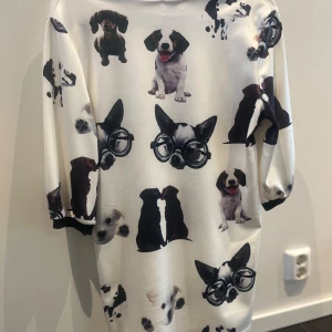 Vit klänning med hundmotiv - Unik vit klänning med ett lekfullt hundmotiv i svart och brunt. Klänningen har trekvartsärmar och en avslappnad passform. Perfekt för hundälskare som vill sticka ut med en rolig och annorlunda stil.